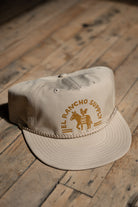A beige baseball cap with a tan embroidered logo reading 'EL Rancho Supply' and a donkey graphic, resting on a rustic wooden plank floor.